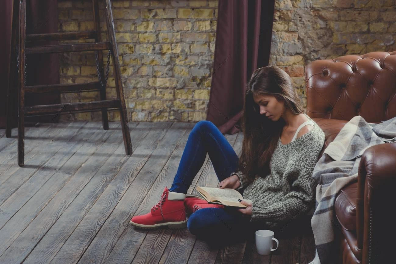 A woman sits on a wooden floor by a leather sofa, reading a book—perfect IG captions moment. She wears a grey sweater, blue jeans, and red shoes. A white mug rests on the floor next to her.