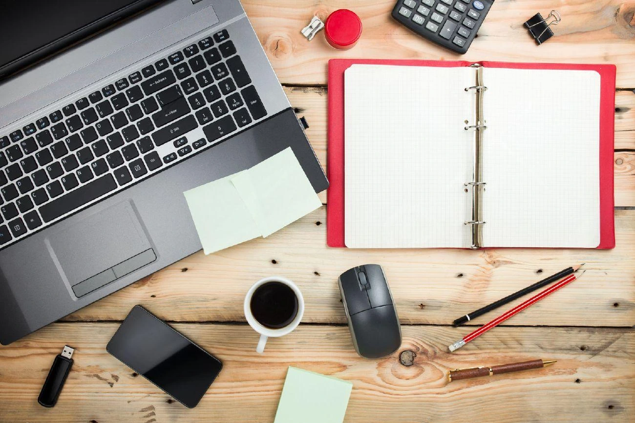 Laptop, open binder, phone, mouse, coffee, pens, sticky notes, calculator, and office supplies arranged on a wooden desk—perfect setup for productivity or crafting creative IG captions.