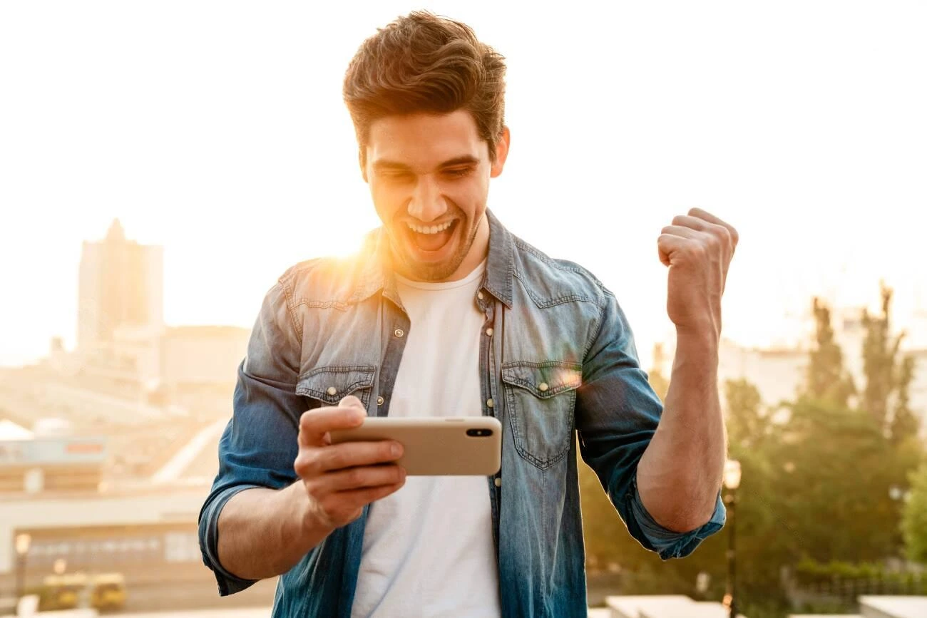 A man outdoors looks at his smartphone, smiling and raising his fist in excitement, as sunlight shines in the background—capturing the perfect moment for IG Captions.