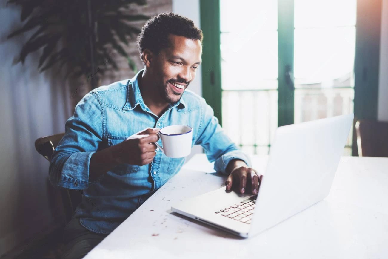 A man in a denim shirt sits at a table, holding a mug and smiling while looking at a laptop in a well-lit room—perfect moment for crafting IG captions.