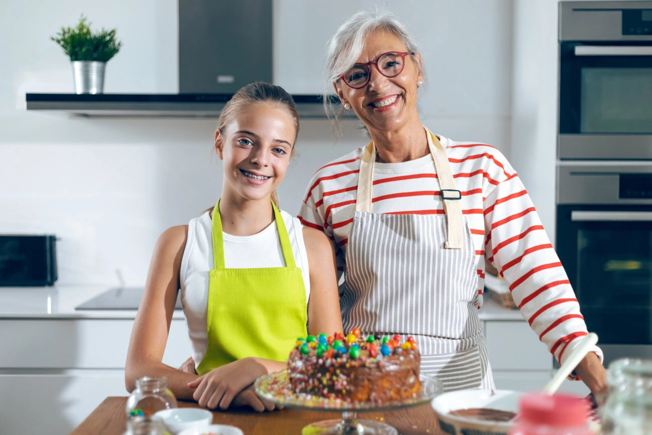 An older woman and a young girl, both wearing aprons, stand in a kitchen smiling behind a decorated chocolate cake on the counter—perfect moment for sweet IG captions.