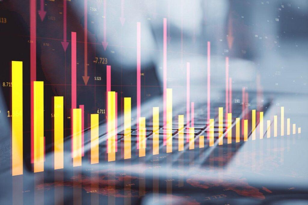 Describe a Picture: Bar and line charts with downward red arrows are superimposed on a blurred image of hands typing on a keyboard, illustrating a decline in financial or market data.